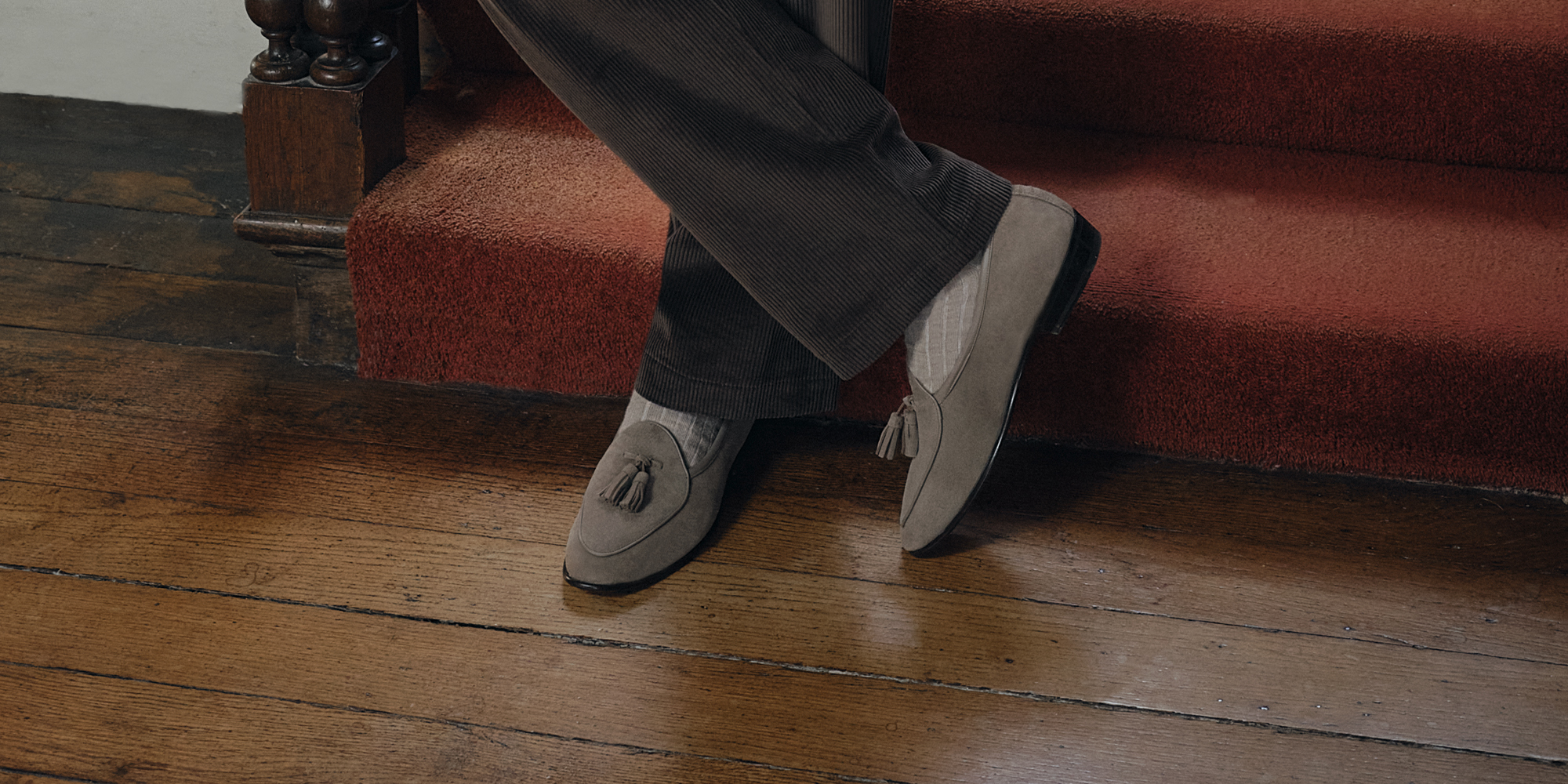 A man wearing greige suede tassel loafers with socks, crossing his legs.